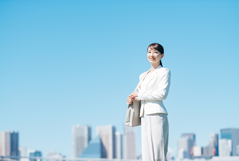 青空の下でにこやかに佇む女性と遠くに見えるオフィス街のイメージ画像